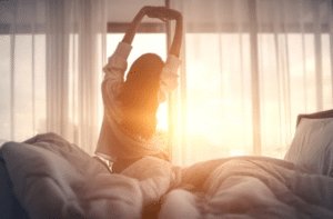 Mulher sentada na cama se espreguiçando, ao fundo o sol esta nascendo por entre as cortinas, ela teve higiene do sono